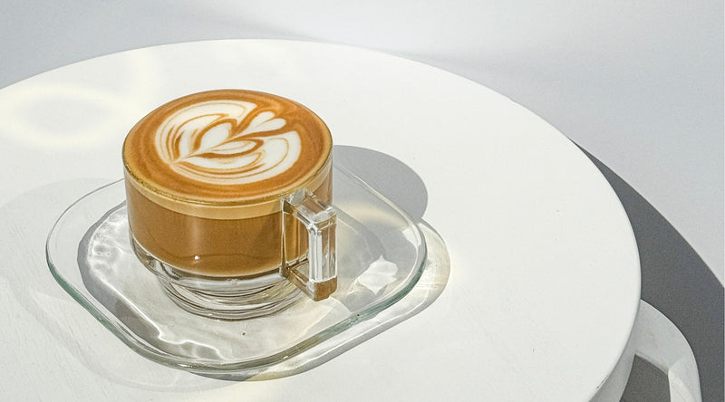  Latte with leaf latte art made using Koatji, served in a clear glass cup on a sunlit white surface.
