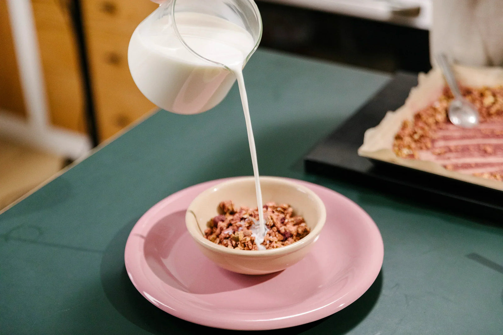 KOATJI milk being poured into a bowl of granola on a pink plate.