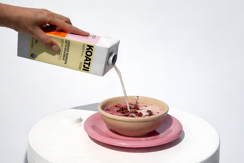 Koatji oat milk being poured from a carton into a pink smoothie bowl on a minimal white table.
