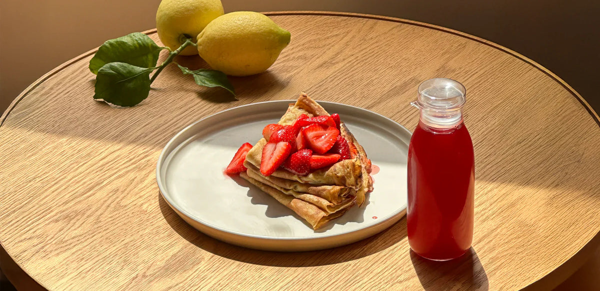 Koatji styled breakfast with strawberry crepes on a ceramic plate and a bottled fruit drink on a sunlit table.