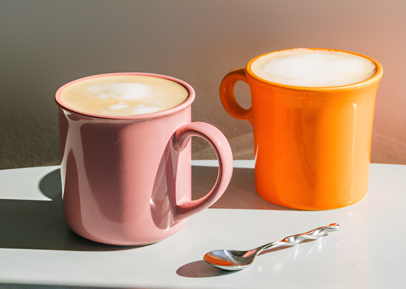 Two mugs of coffee with KOATJI plant milk on a table with soft lighting and a spoon on the side.